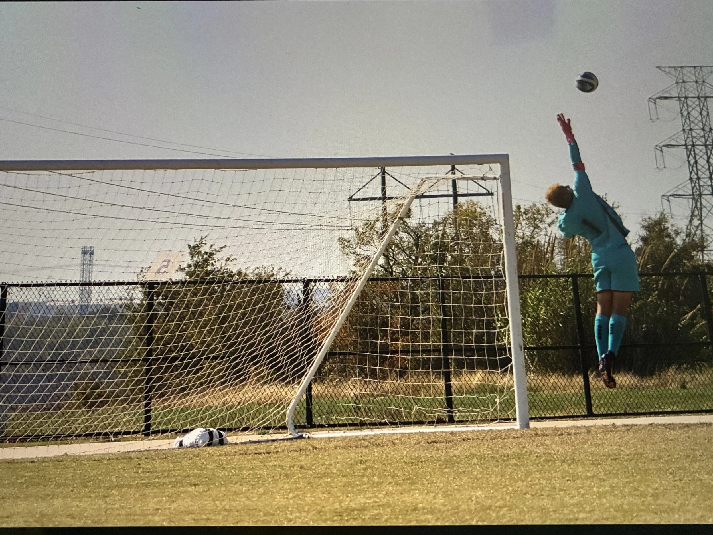A soccer goalkeeper in a blue uniform jumps to catch a soccer ball near the goalpost on a grassy field, with a soccer ball on the ground and trees in the background.