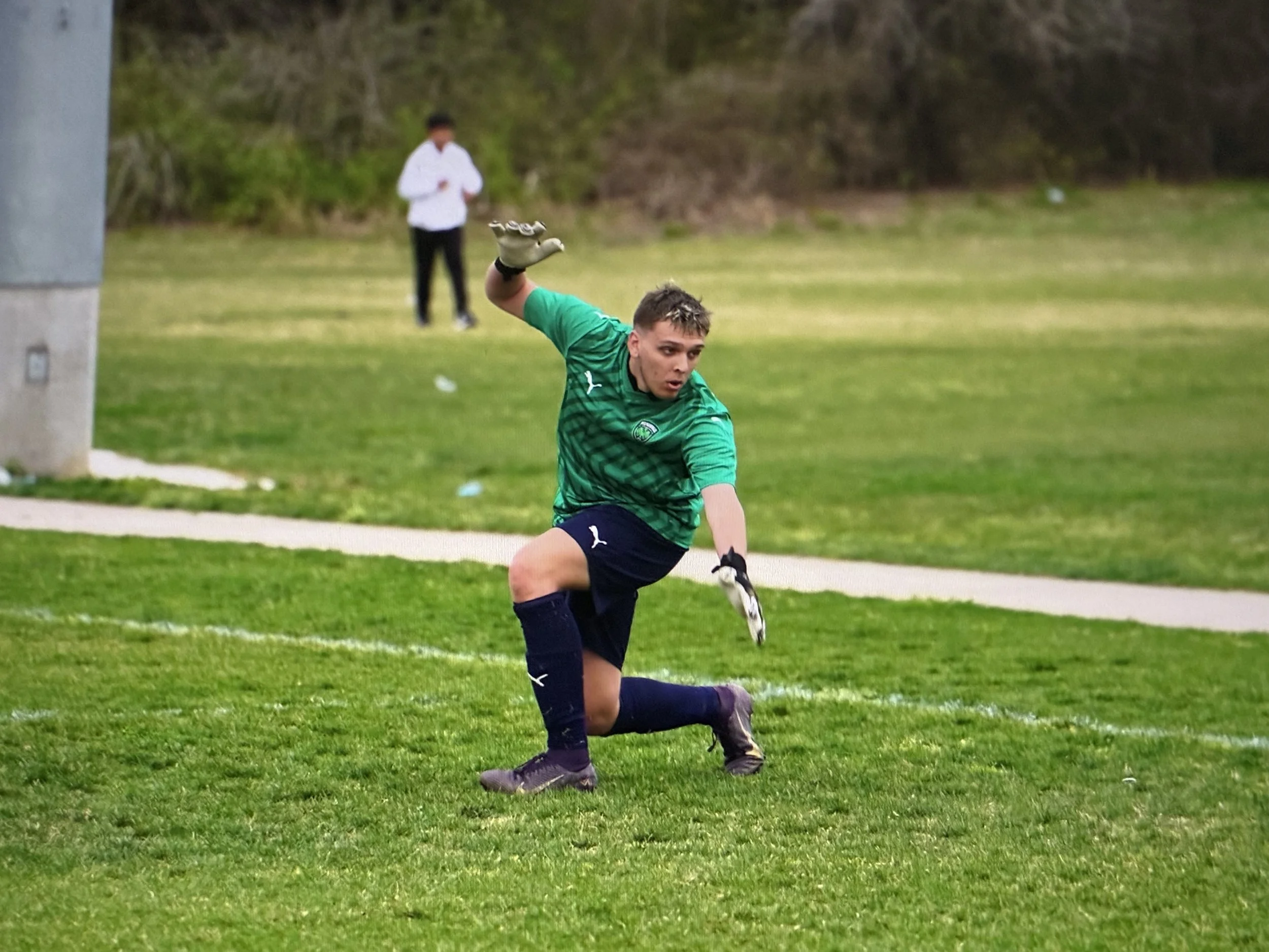 A young male soccer goalkeeper in a green jersey and navy shorts dives to catch a soccer ball on a grassy field during daytime. A person in a white jacket stands in the background near a wooded area.