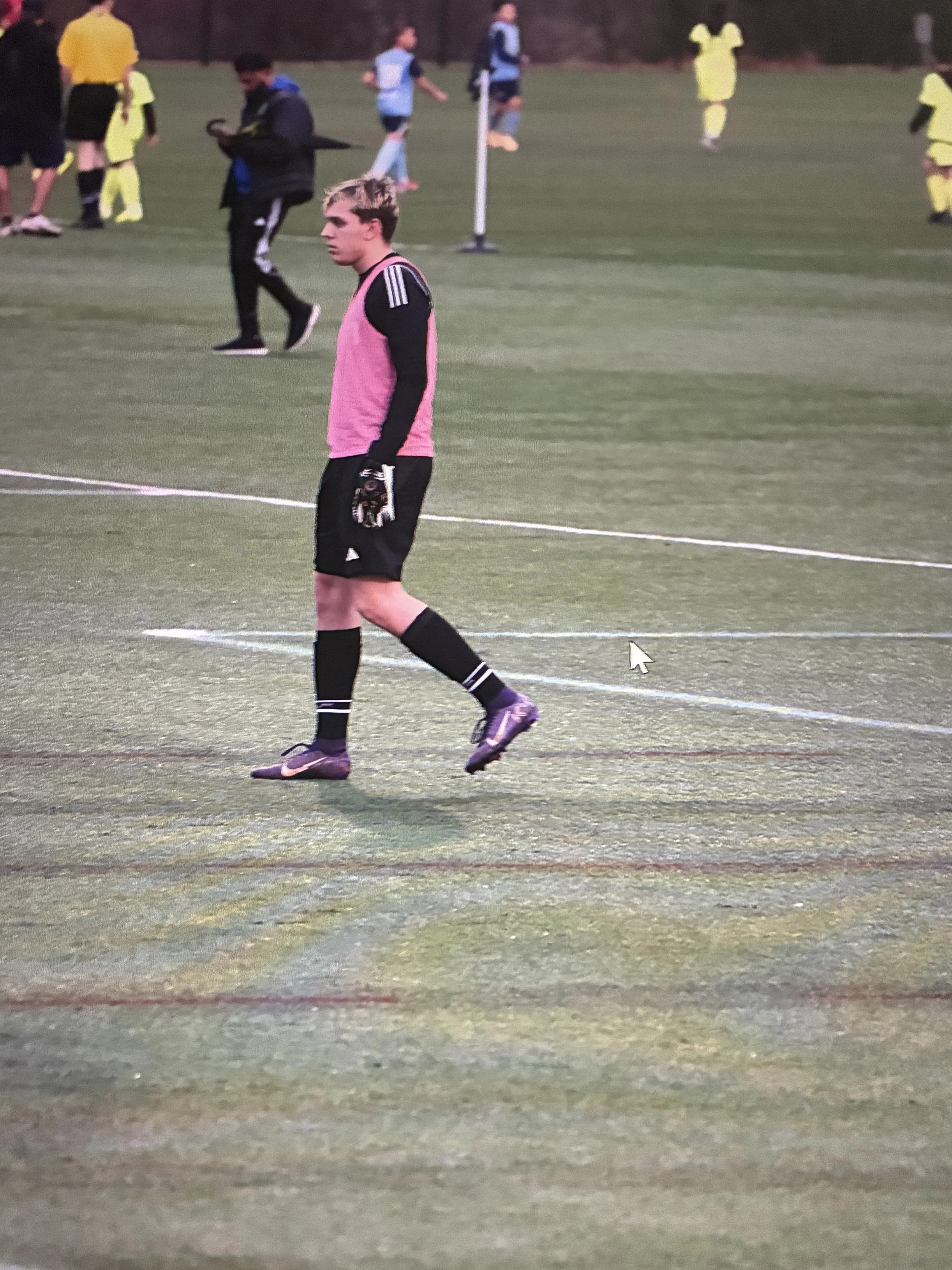 A young male soccer goalkeeper wearing a pink and black uniform standing on a soccer field, with other players and a referee in the background.