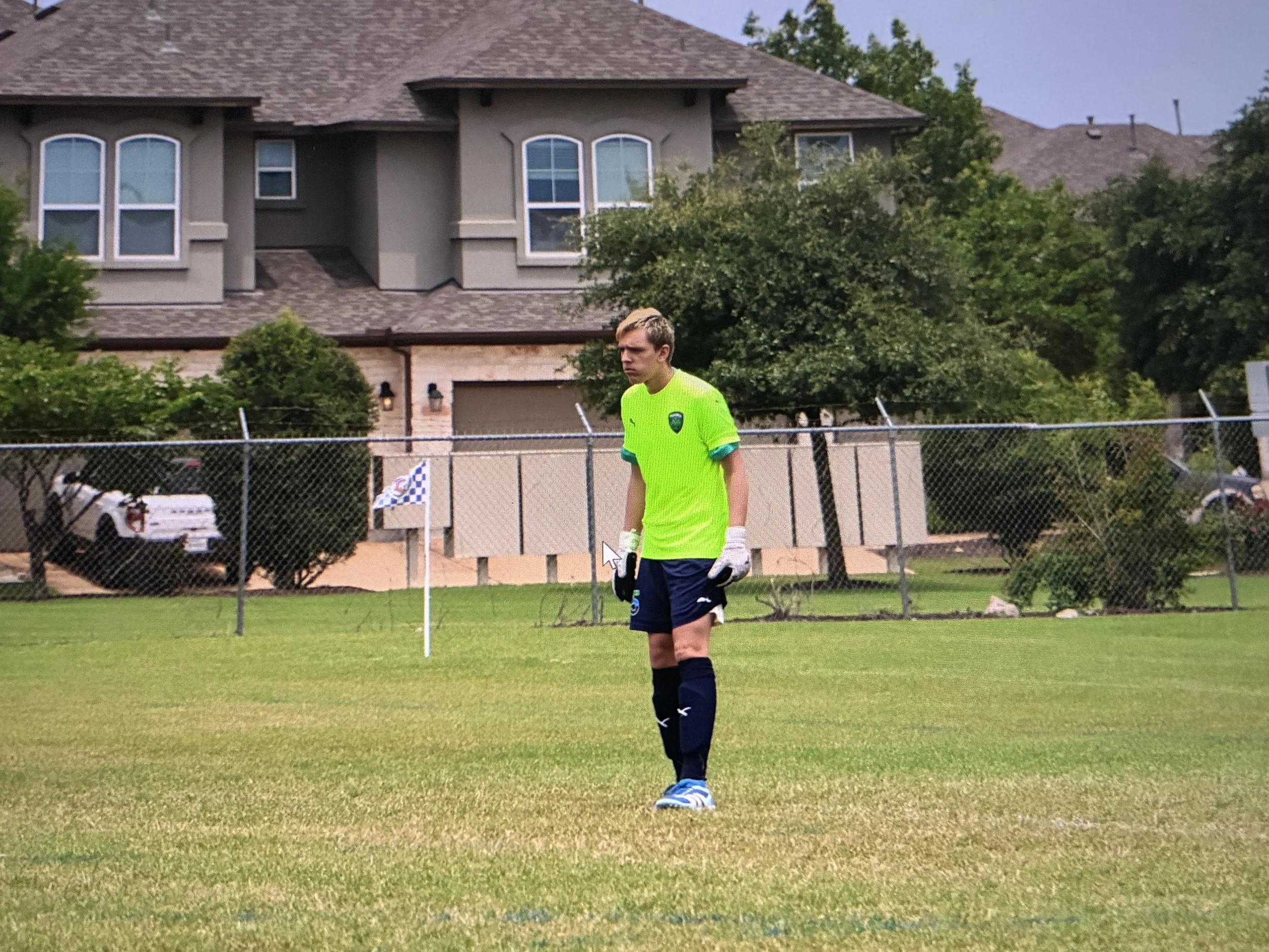 A young male soccer player with blonde hair, wearing a neon yellow jersey, dark shorts, black socks, blue and white cleats, and goalkeeper gloves, standing on a grassy field during daytime. There is a chain-link fence with sideline markers, and suburban houses with trees in the background.