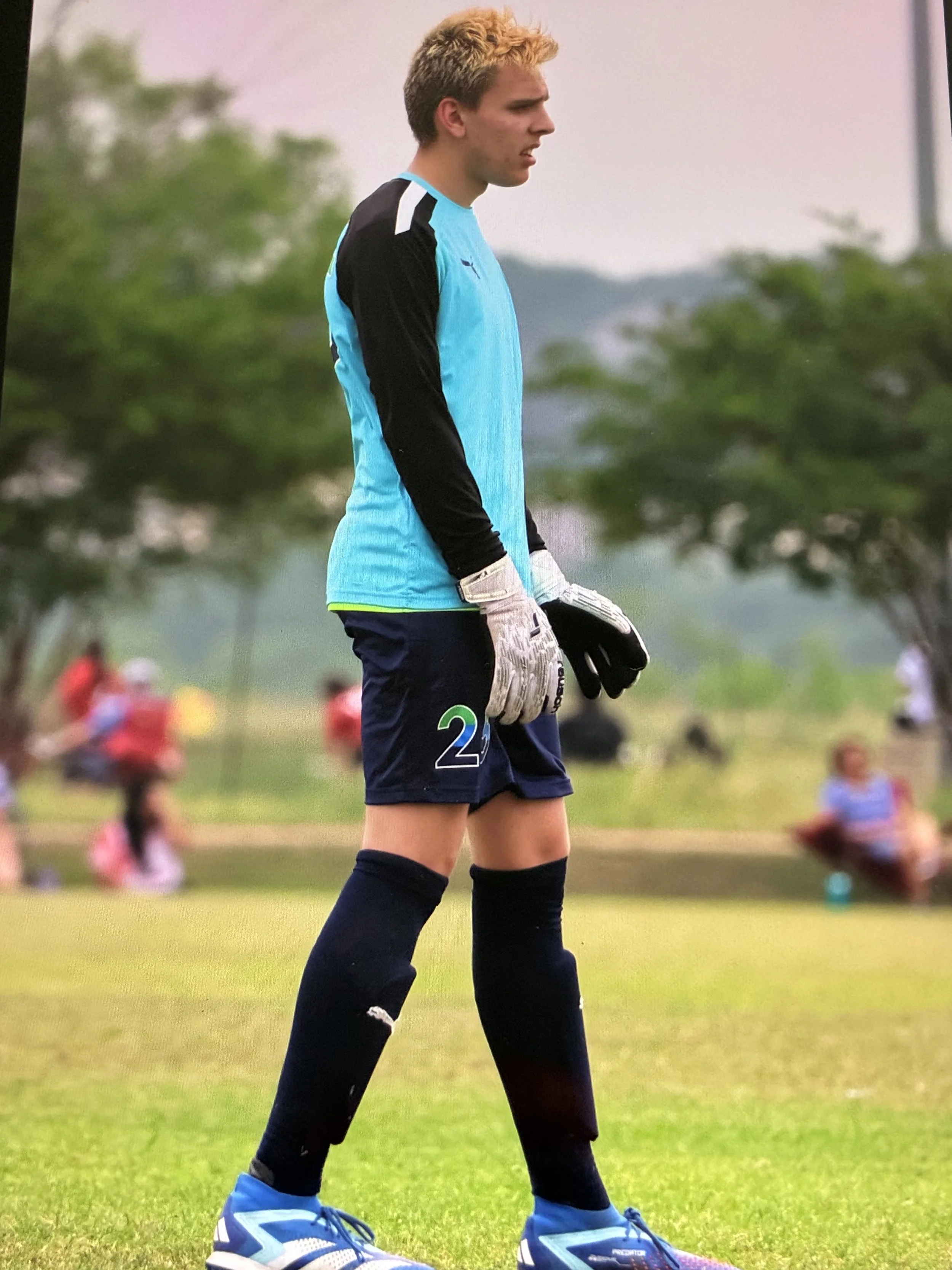 A young male soccer goalkeeper wearing a light blue jersey with black sleeves, black shorts, black knee-high socks, and blue cleats, standing on a grassy field with trees and blurred spectators in the background.