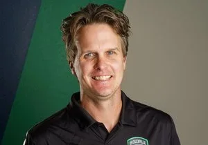 Portrait of a smiling man with wavy brown hair wearing a black collared shirt against a background with green and gray sections.