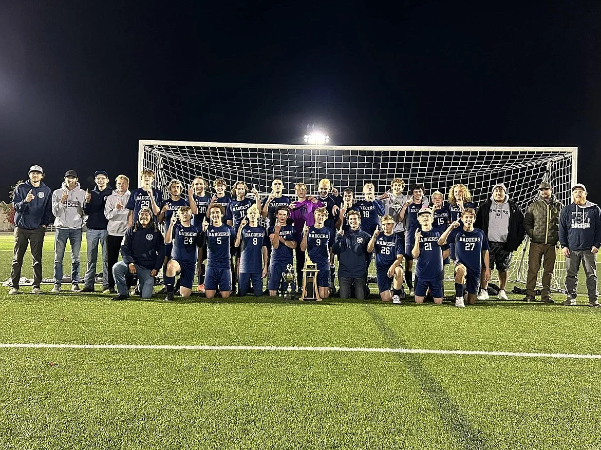 A group of soccer players and coaches standing and kneeling in front of a goalpost on a soccer field at night, celebrating a victory with trophies.