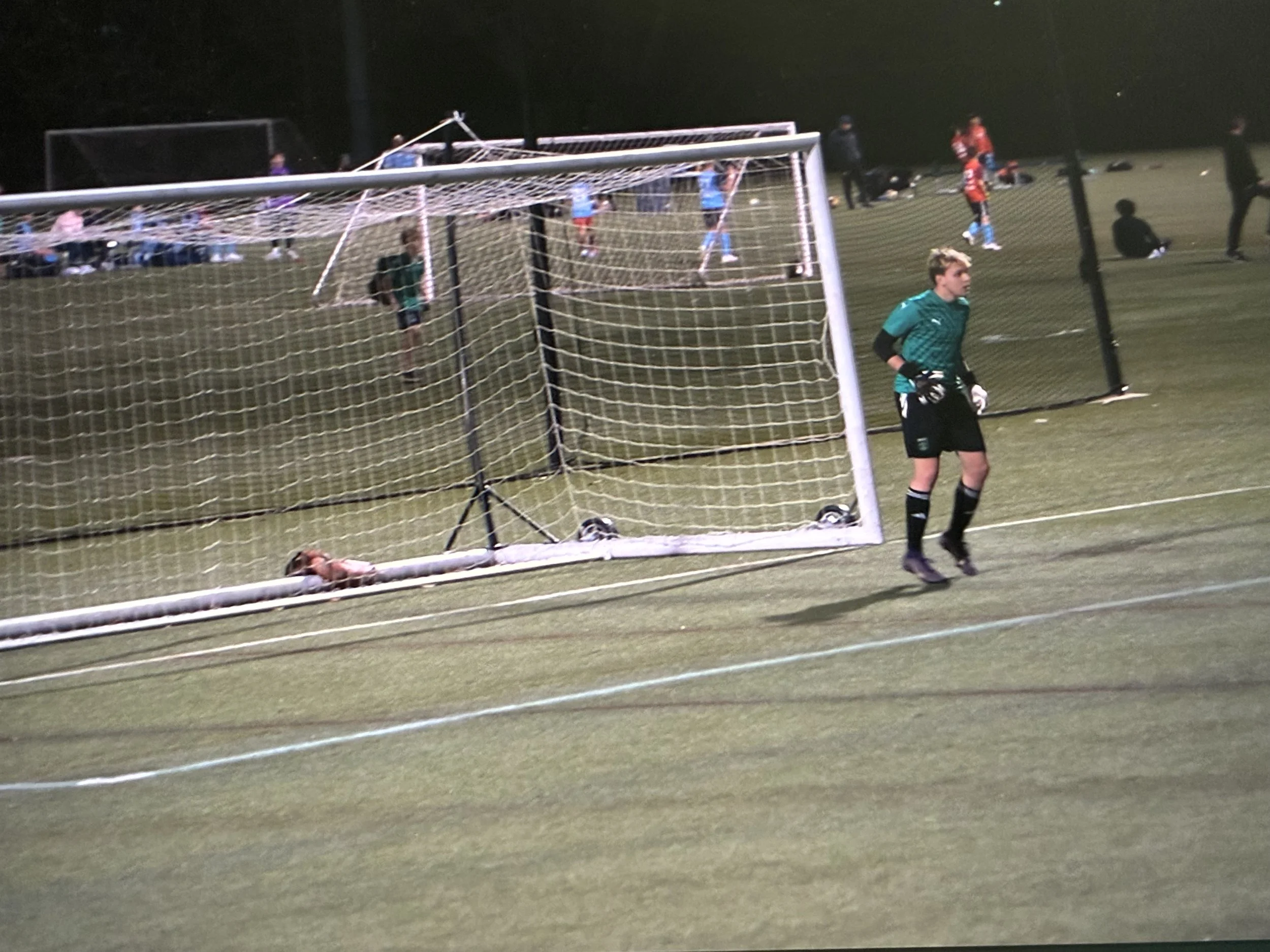 A soccer field at night with players, a goal, and a goalkeeper in green standing in front of the goal. A player in red jersey near the goal is on the ground, possibly having fallen or injured.
