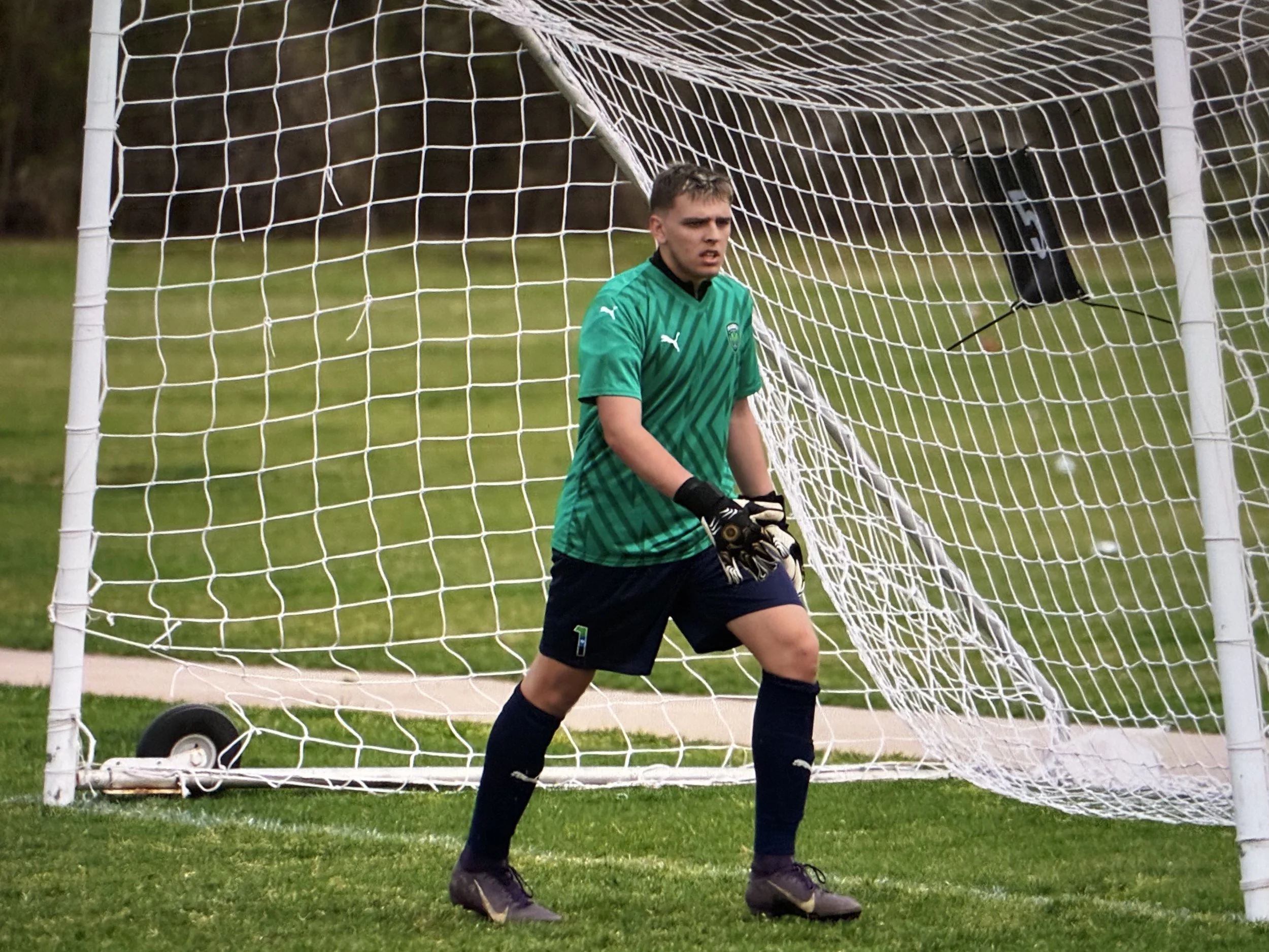 A male soccer goalkeeper standing inside the goal with a serious expression, wearing a green jersey, black shorts, gloves, and black socks, with a soccer ball on the grass in front of him.