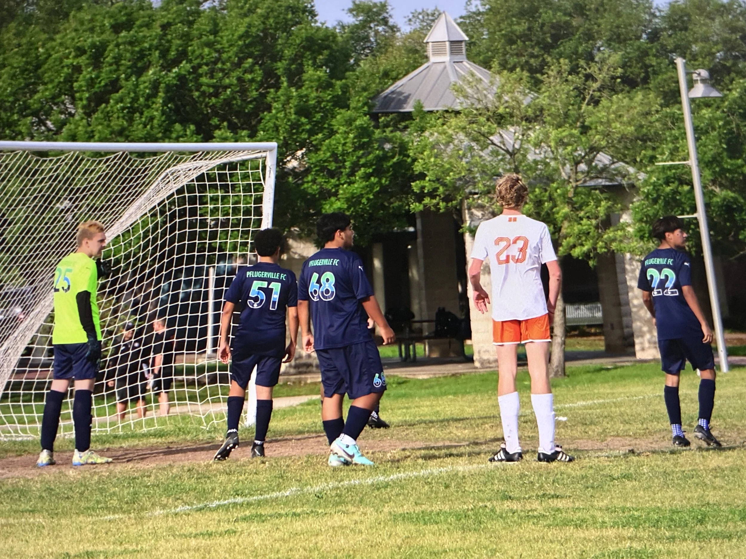 Soccer players standing near the goalpost on a grassy field during a game, with a goalkeeper in a neon green jersey and others wearing blue and white jerseys with the numbers 51, 68, and 23 visible, and trees in the background.