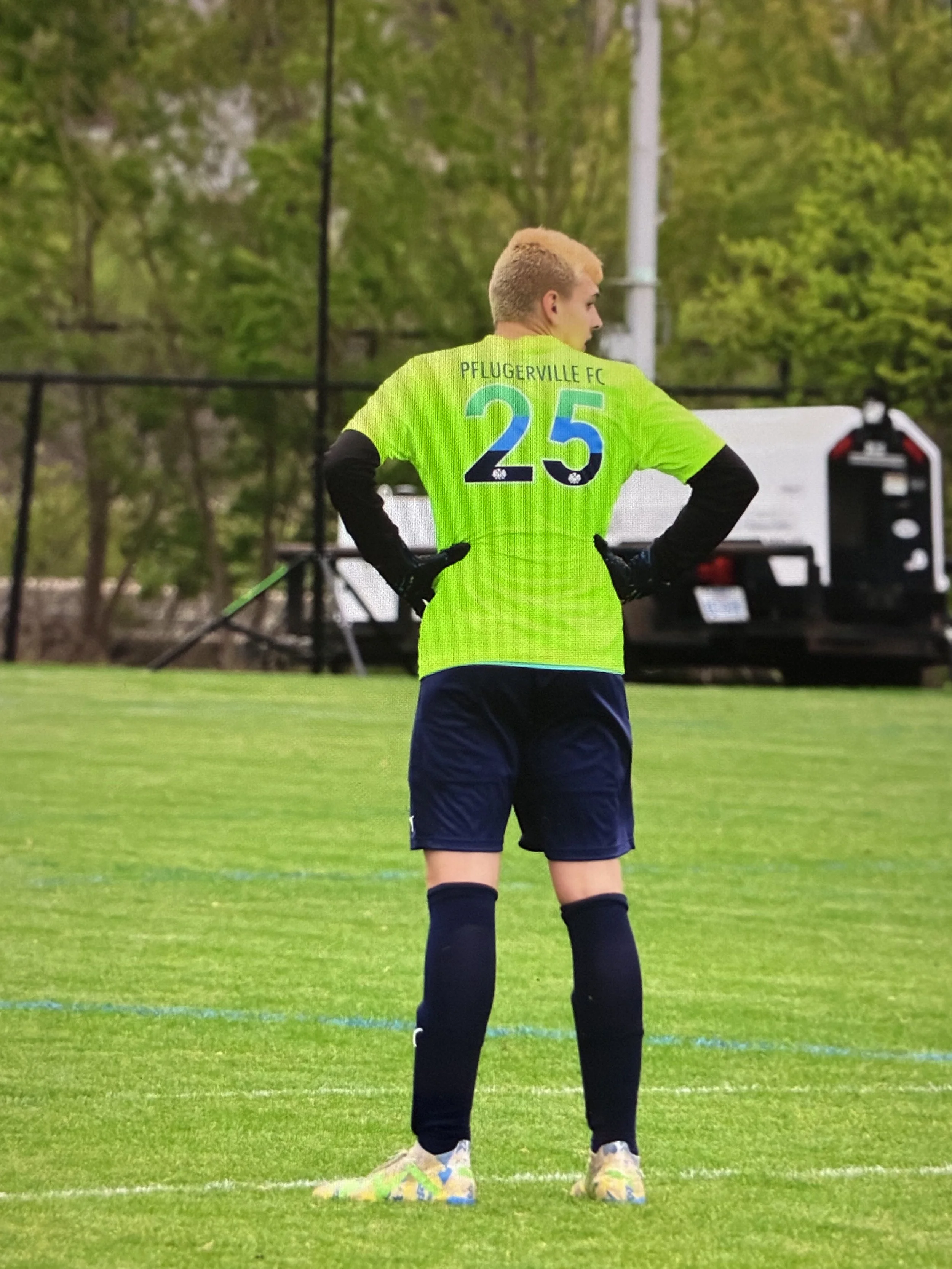 A soccer player wearing a yellow jersey with the number 25 and 'Pflugerville FC' written on the back, standing on a grassy field, with trees and equipment visible in the background.