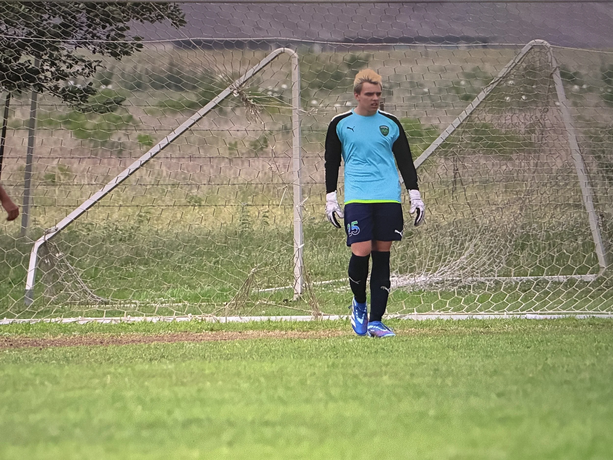 A young male soccer goalkeeper standing in front of a damaged soccer goal net on a grassy field, wearing a blue goalkeeper jersey with black sleeves, black shorts, and blue shoes, with a determined expression.