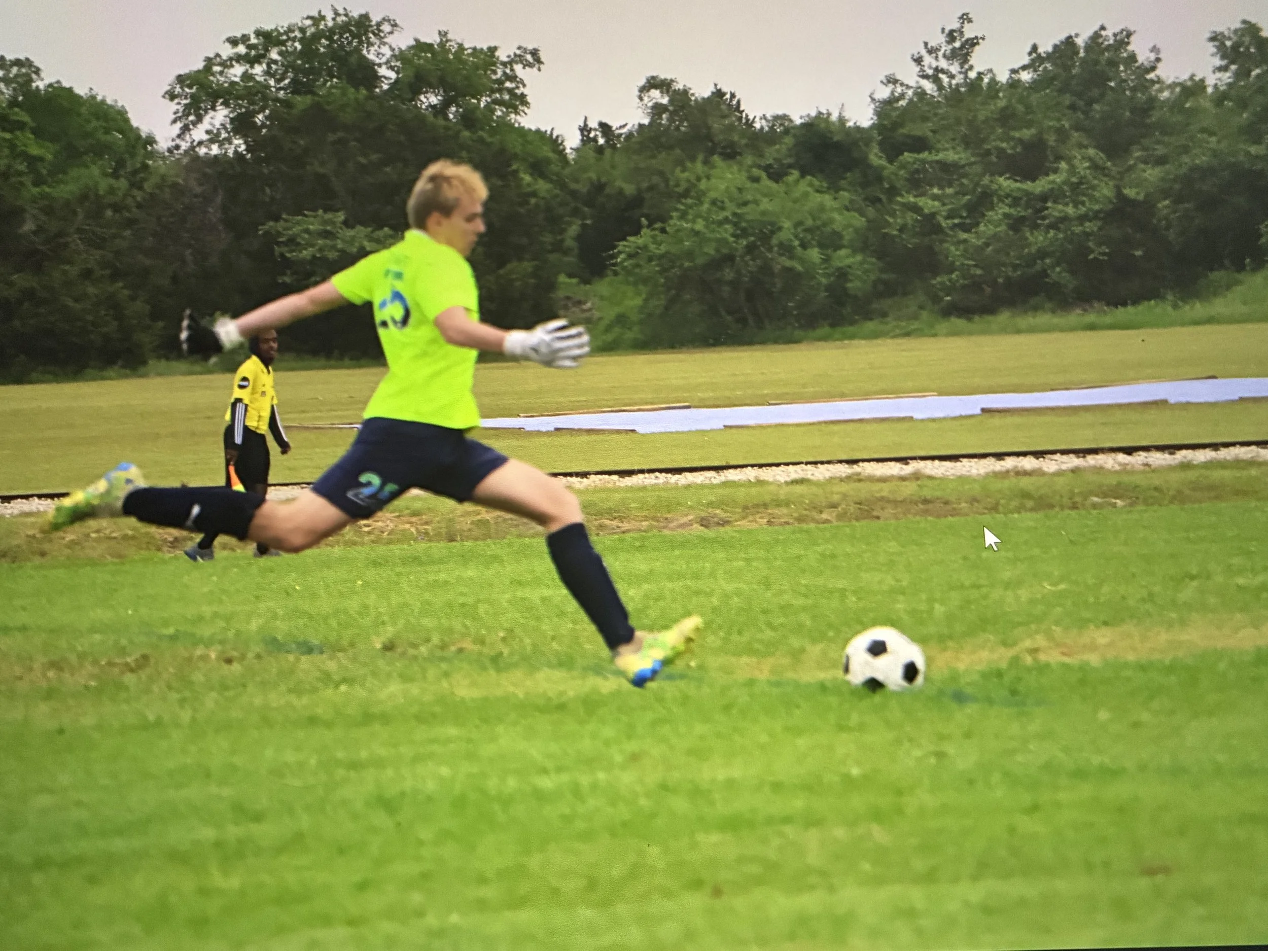 A young man in a bright green soccer jersey and black shorts is running on a soccer field with a soccer ball in front of him. There is a second person in a yellow and black uniform standing behind him on the field. In the background, there are trees and a running track.