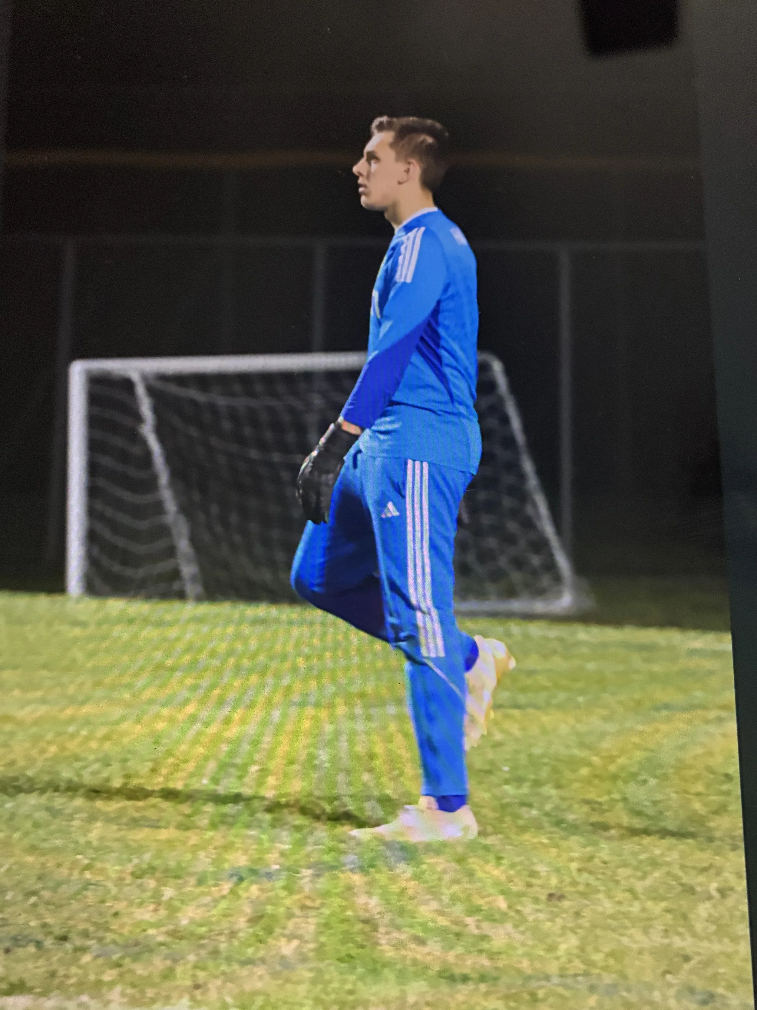A young man wearing a blue athletic outfit, including a long-sleeve shirt and pants with white stripes, is standing on a grassy field near a soccer goalpost at night, with his hands in his pockets and looking to his left.