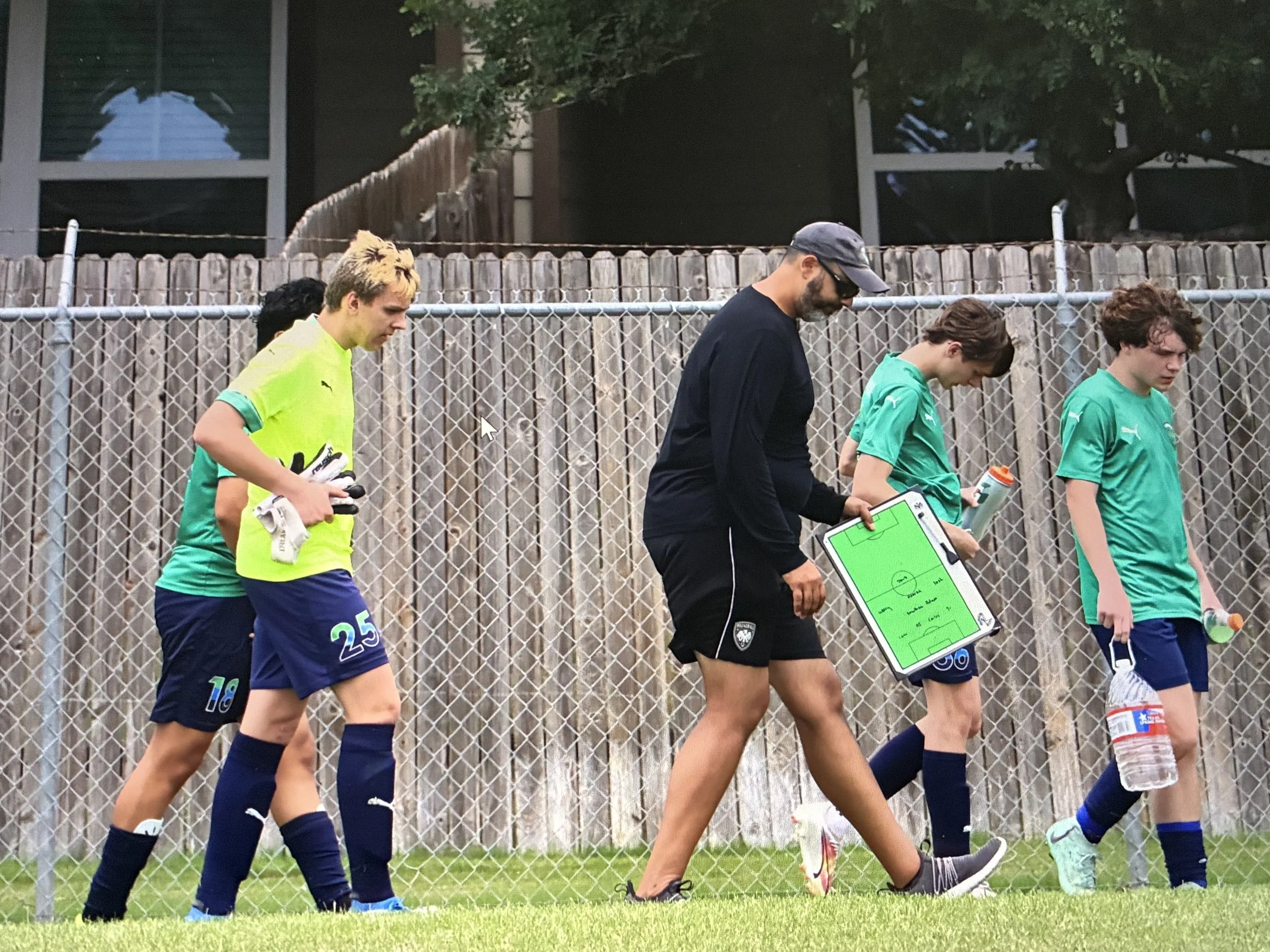 A group of young soccer players walking along a fence, a coach holding a clipboard, and one player holding a water bottle, with a building and trees in the background.