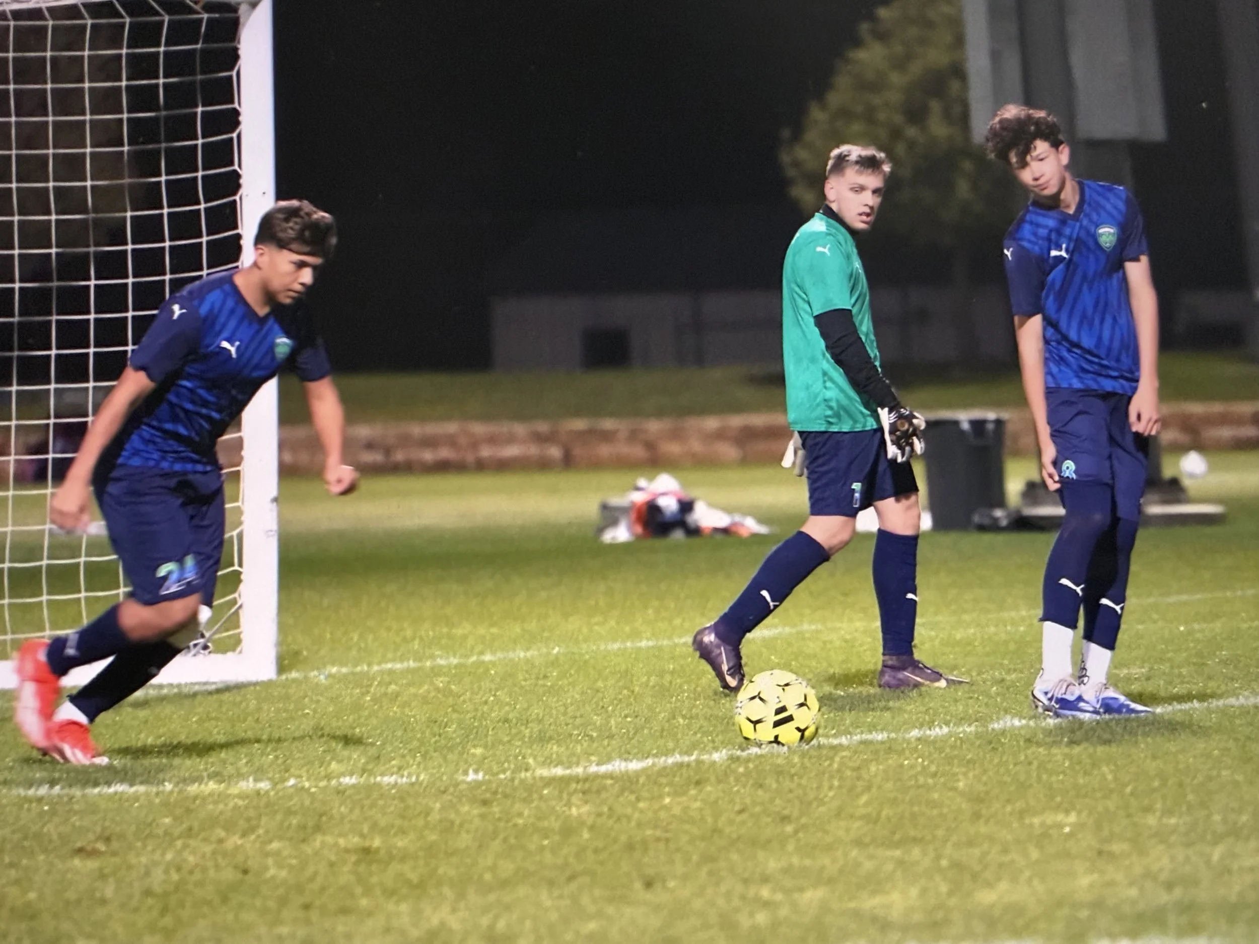 Three young male soccer players standing on a field at night, with one about to kick a yellow and black patterned soccer ball; two of them are wearing blue jerseys, and one in the middle is dressed in a green goalkeeper jersey. A soccer goal with netting is visible on the left.