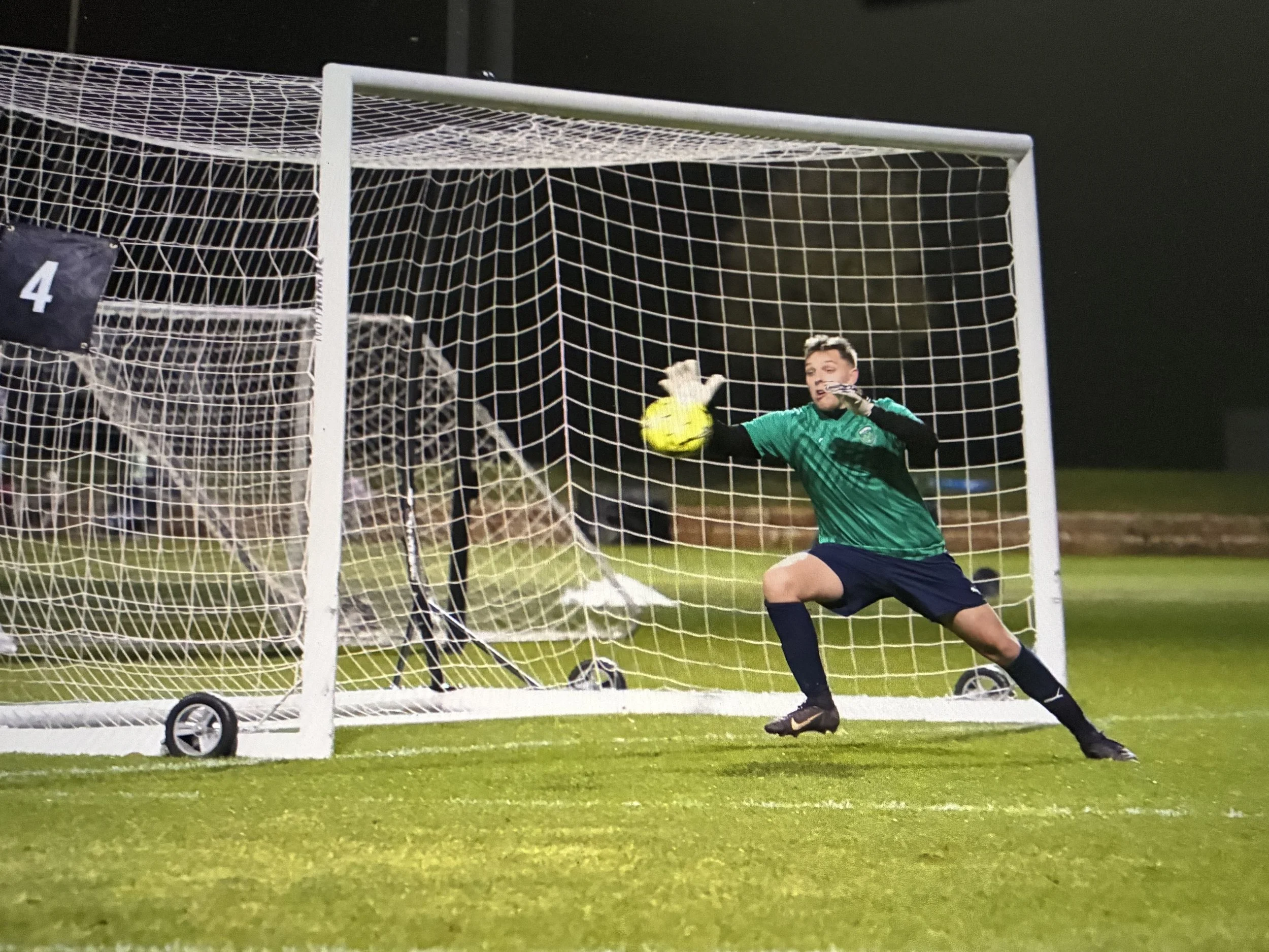 A soccer goalkeeper in a green jersey and black shorts making a save during a game at night. The goalkeeper is reaching out with gloved hands to catch a bright yellow soccer ball just inside the goal, with a white goalpost and net behind him and a dark evening sky overhead.