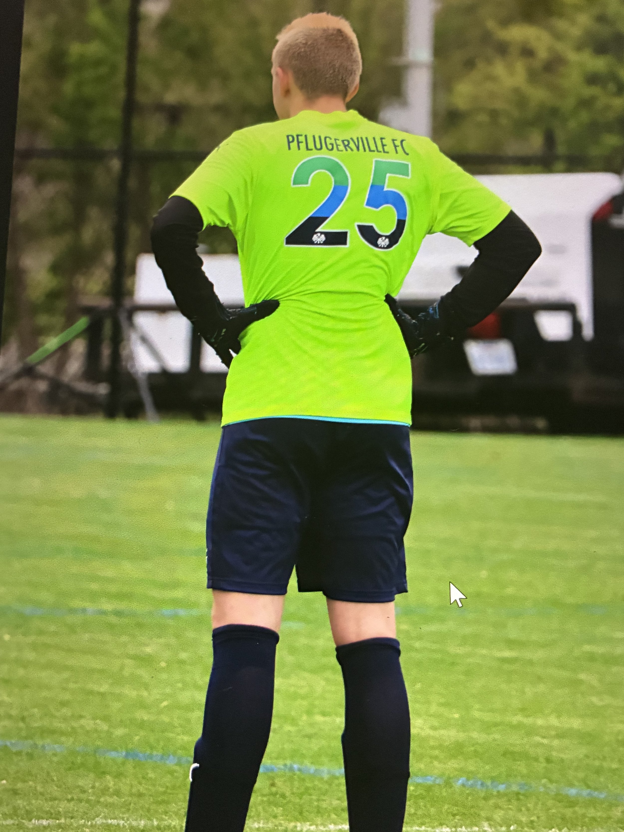 Back view of a soccer player wearing a bright yellow green jersey with the number 25 and 'Pflugerville FC' written on it, standing on the field with hands on hips.