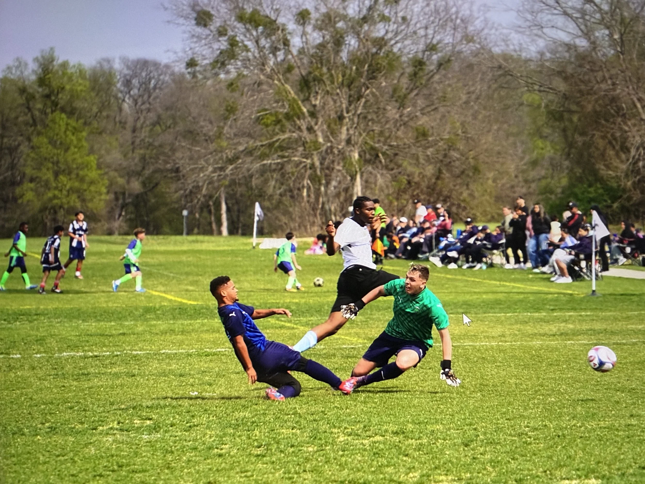 A soccer match with players on the field, featuring a player sliding to kick the ball, another player in a green goalkeeper uniform on the ground, and spectators in the background watching the game in a park with trees.