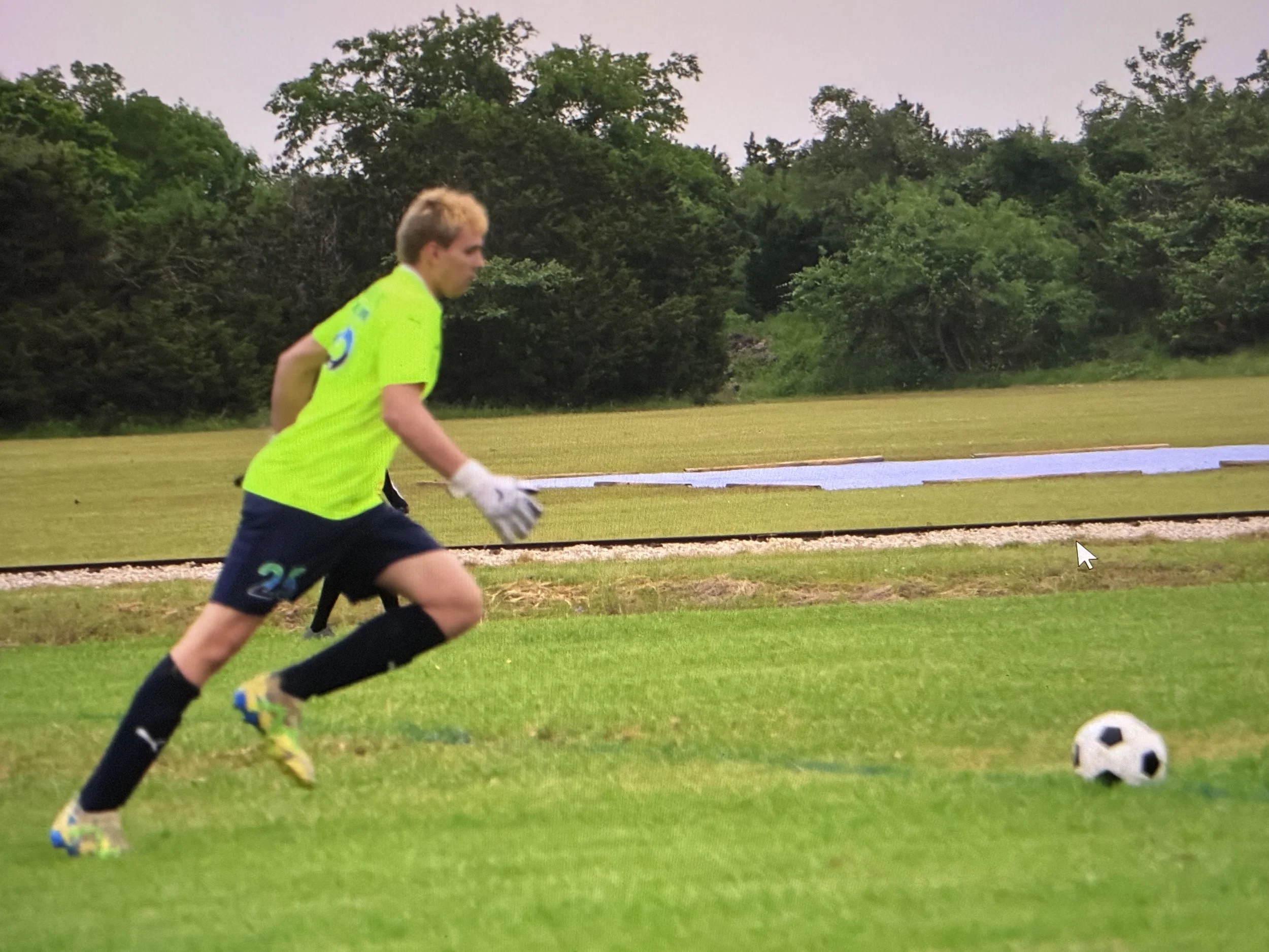 A young male soccer player in a neon yellow jersey and black shorts runs on a grassy field with a soccer ball nearby. Trees and a running track are visible in the background.