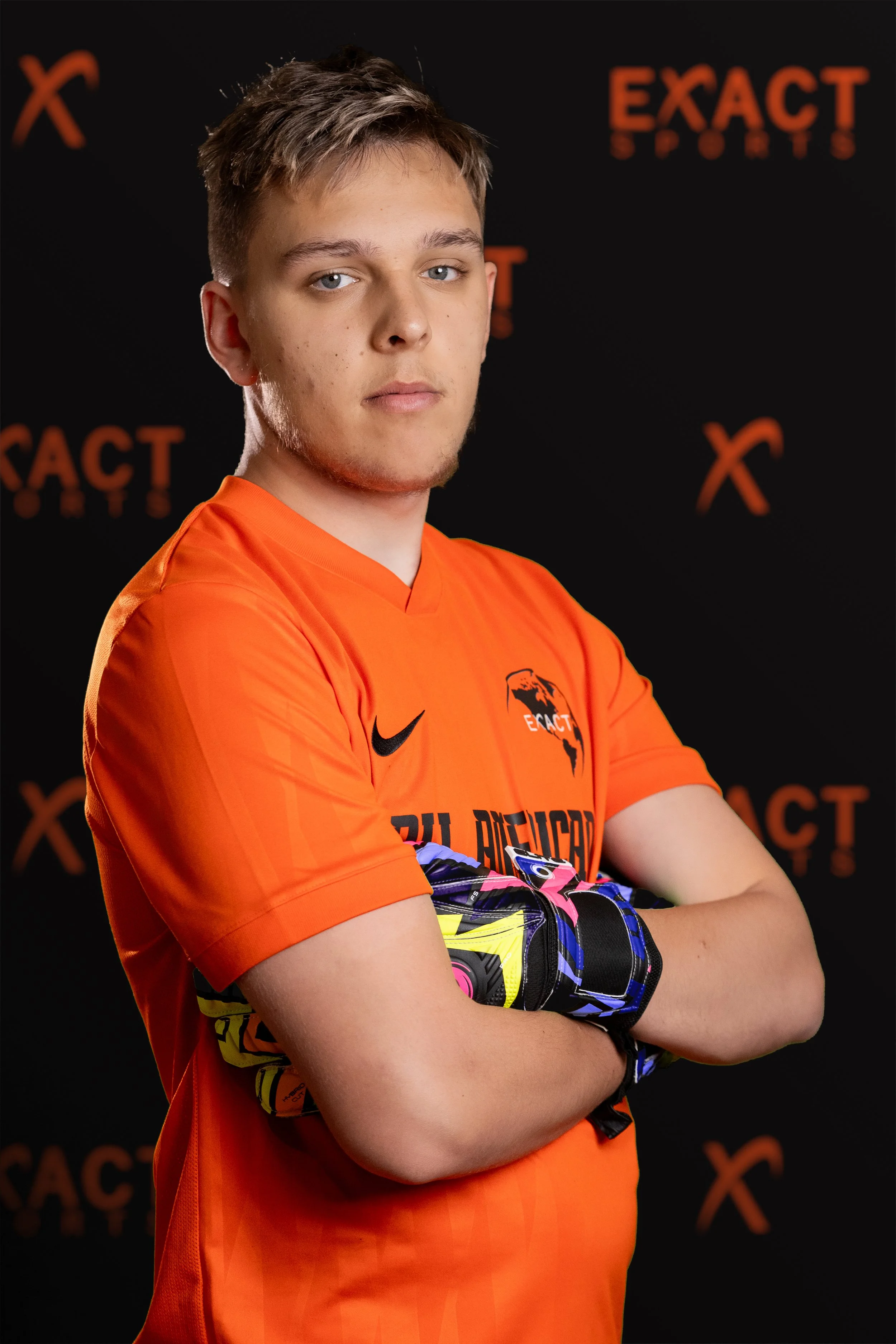 A young male athlete wearing an orange sports jersey and colorful gloves, standing with arms crossed in front of a black background with the 'Exact Sports' logo.