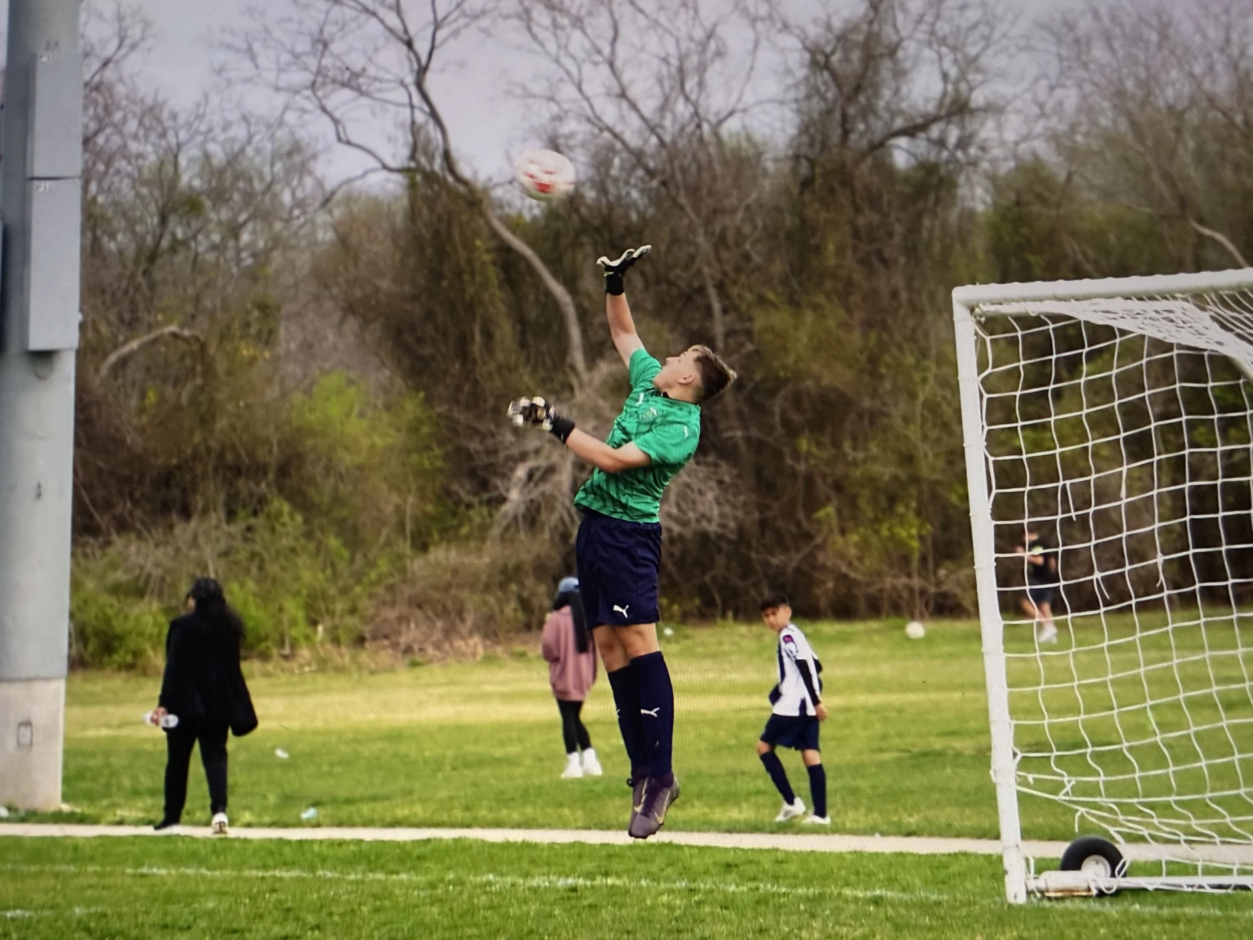 A young male soccer goalkeeper in a green jersey and black shorts is jumping with his arm extended to catch a soccer ball in front of the goalpost on a grassy field. Several children and a woman are in the background, with trees and cloudy sky behind them.