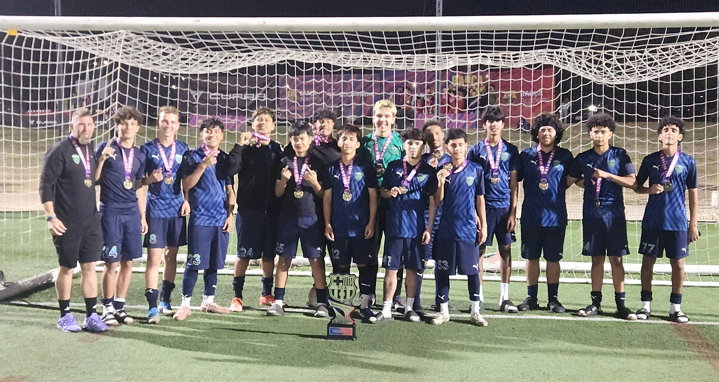 A youth soccer team in blue uniforms with medals around their necks, standing in front of a goal on a field at night, celebrating their victory with a trophy and plaque.