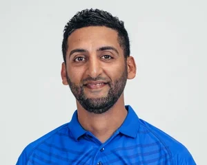 A man with a beard and short dark hair wearing a blue polo shirt, smiling at the camera against a plain light background.