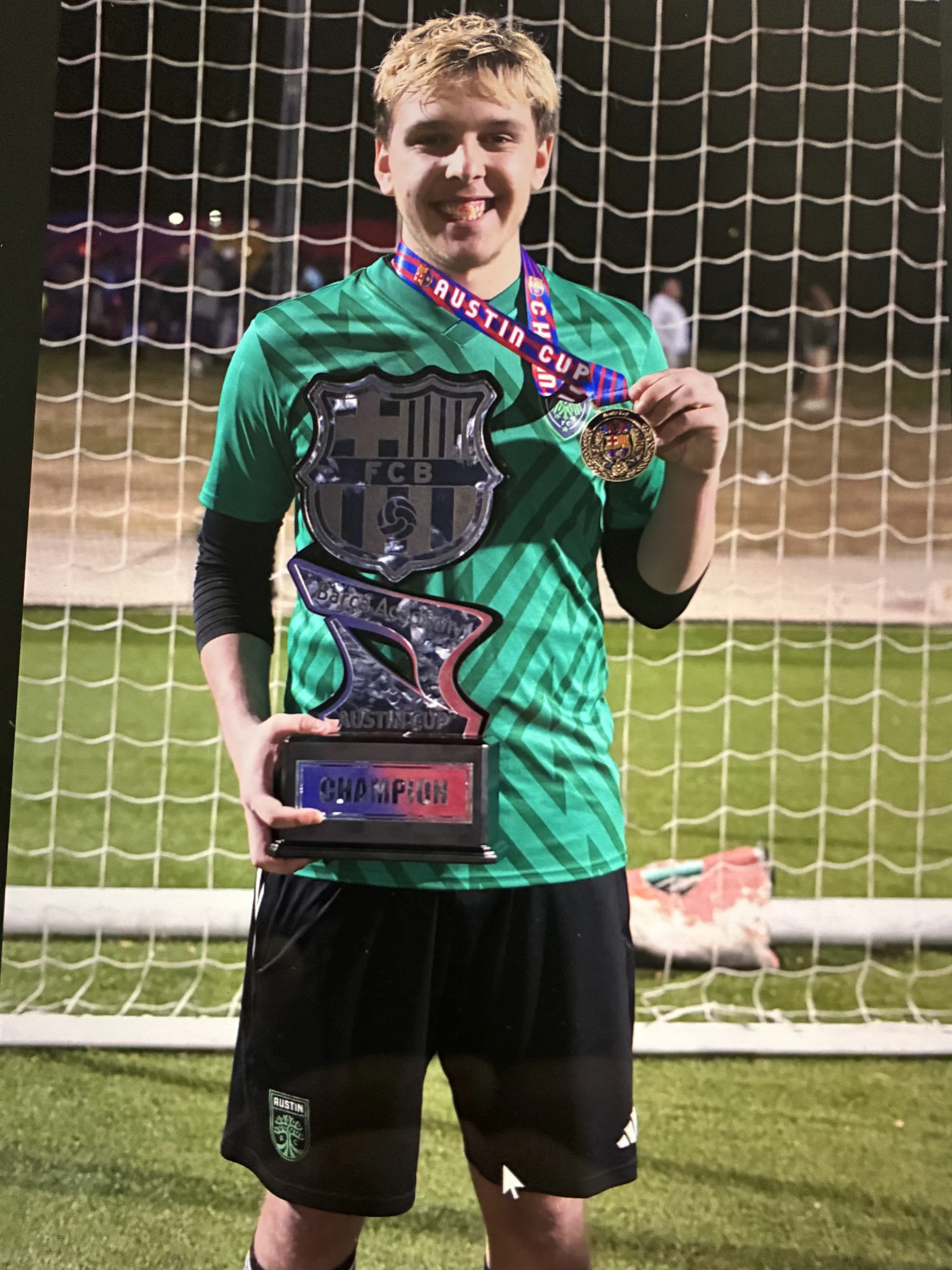 A young male soccer player standing on a field at night, holding a trophy and wearing a medal around his neck. He is smiling with his tongue slightly out and is dressed in a green athletic jersey, black shorts, and black socks. The background features a soccer goal net and some blurred people.