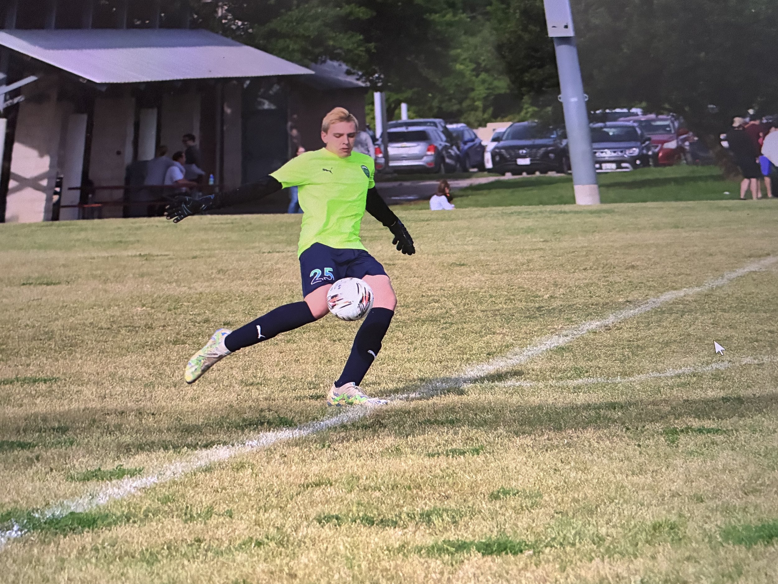 A young man wearing a neon green shirt and black gloves, playing soccer on a grass field during dusk. He is wearing navy blue shorts with the number 25, and white cleats with colorful accents. There are parked cars, trees, and a few people in the background.