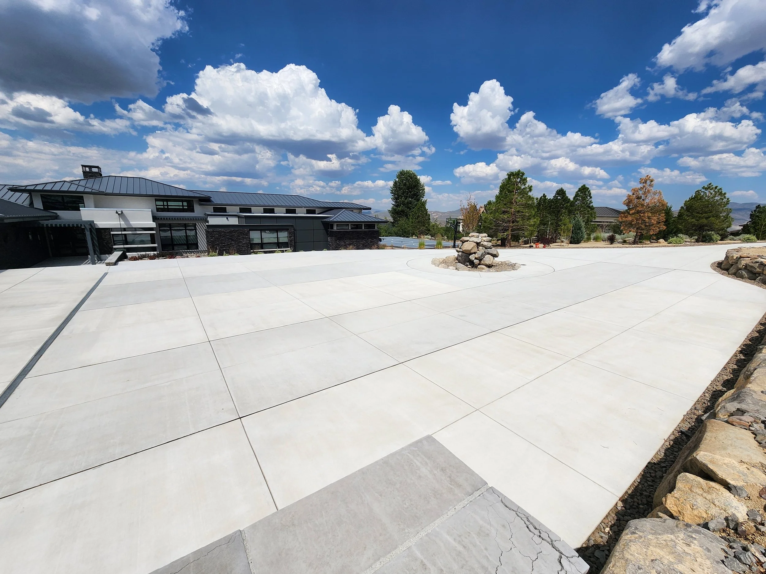 Large, smooth concrete driveway leading to a modern house with a metal roof, surrounded by trees and a partly cloudy blue sky.