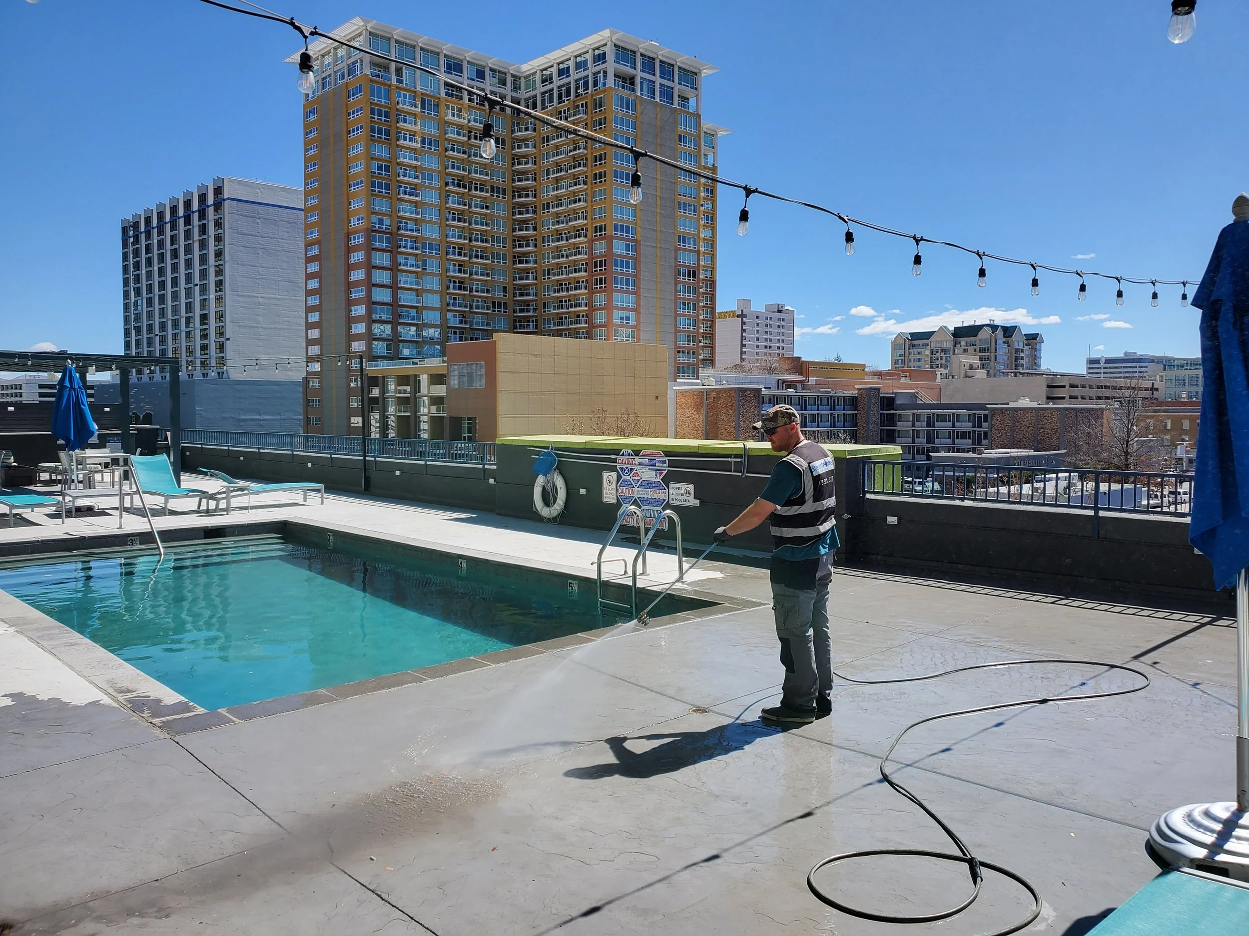 A man cleaning a rooftop swimming pool with a hose on a sunny day, surrounded by lounge chairs and string lights, with tall buildings in the background.