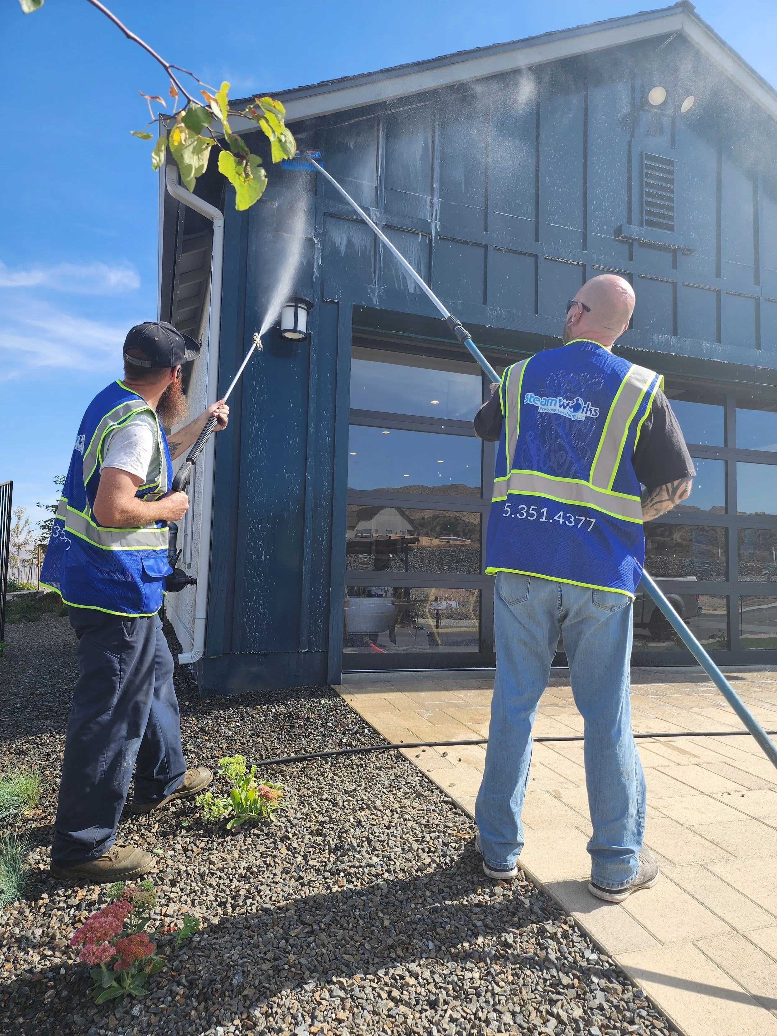 Two workers in high-visibility safety vests pressure washing the outside of a building with a large window under a clear blue sky.
