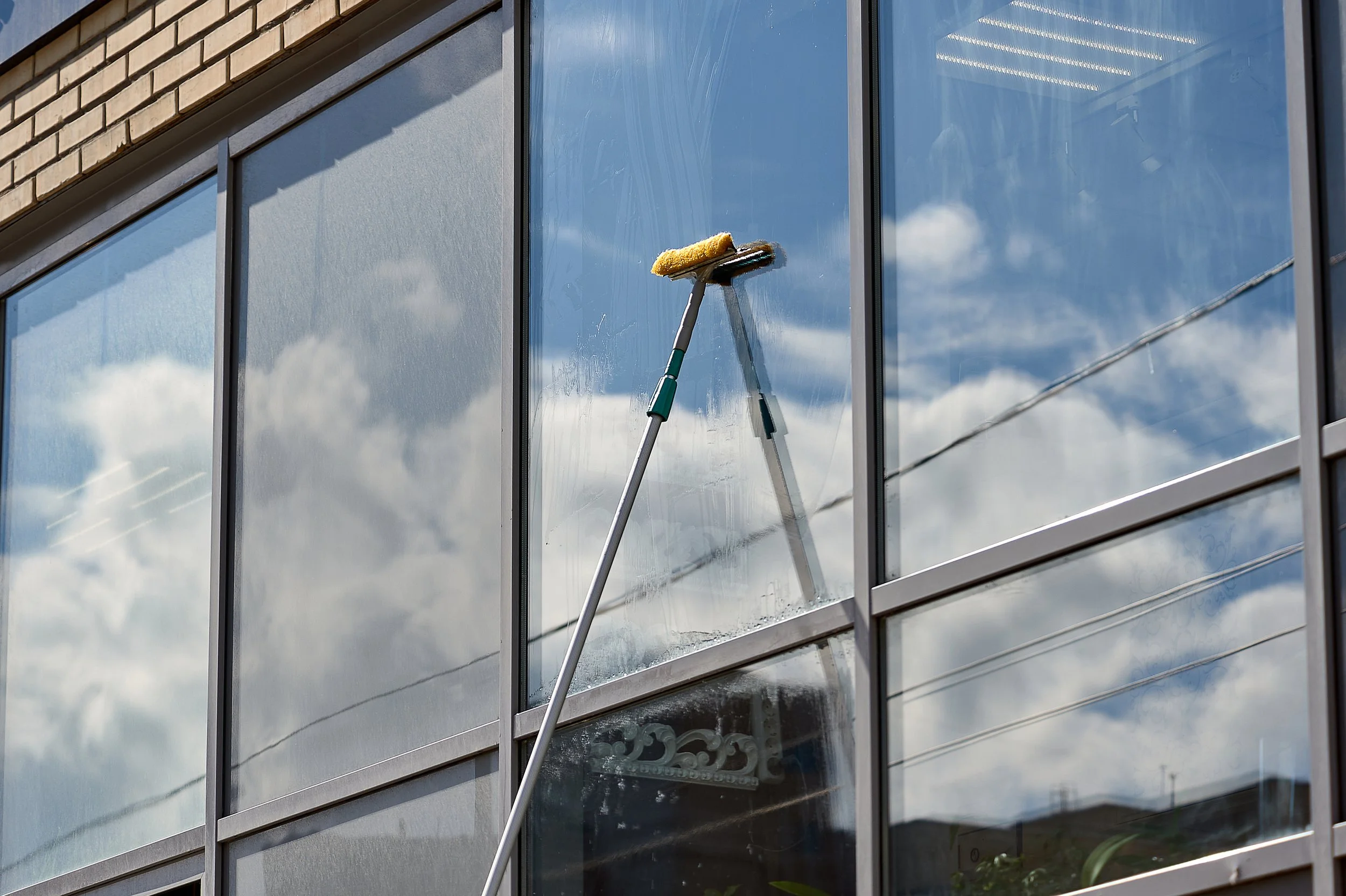 A window washer's squeegee with a yellow cleaning sponge attached to it on a tall pole is cleaning the exterior glass window of a building. The window reflects the cloudy sky.