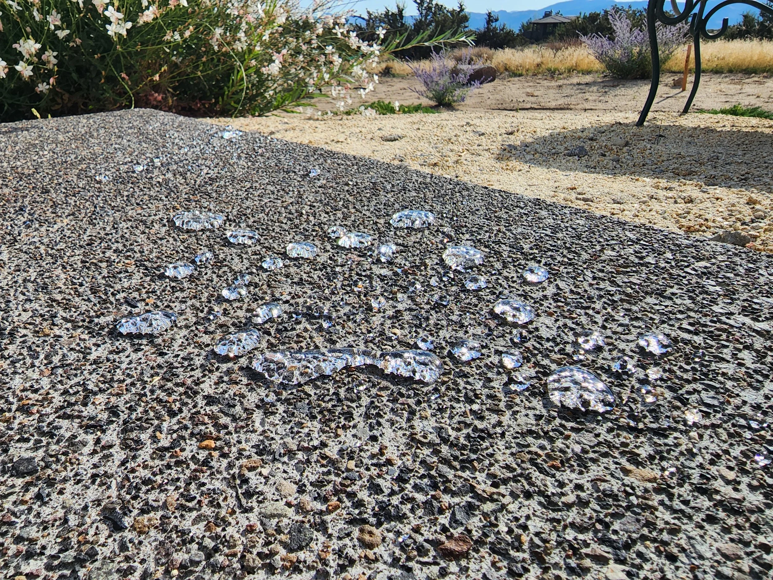 Close-up of several water droplets on a textured gray stone surface outdoors, with plants and a chair in the background.