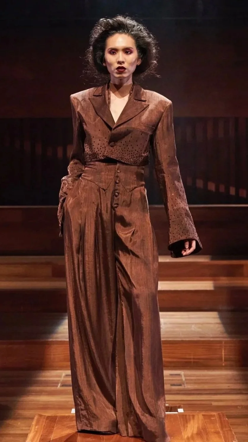 A woman standing on stage wearing a vintage brown suit with wide-leg trousers and a cropped blazer, with dark, curled hair and makeup.