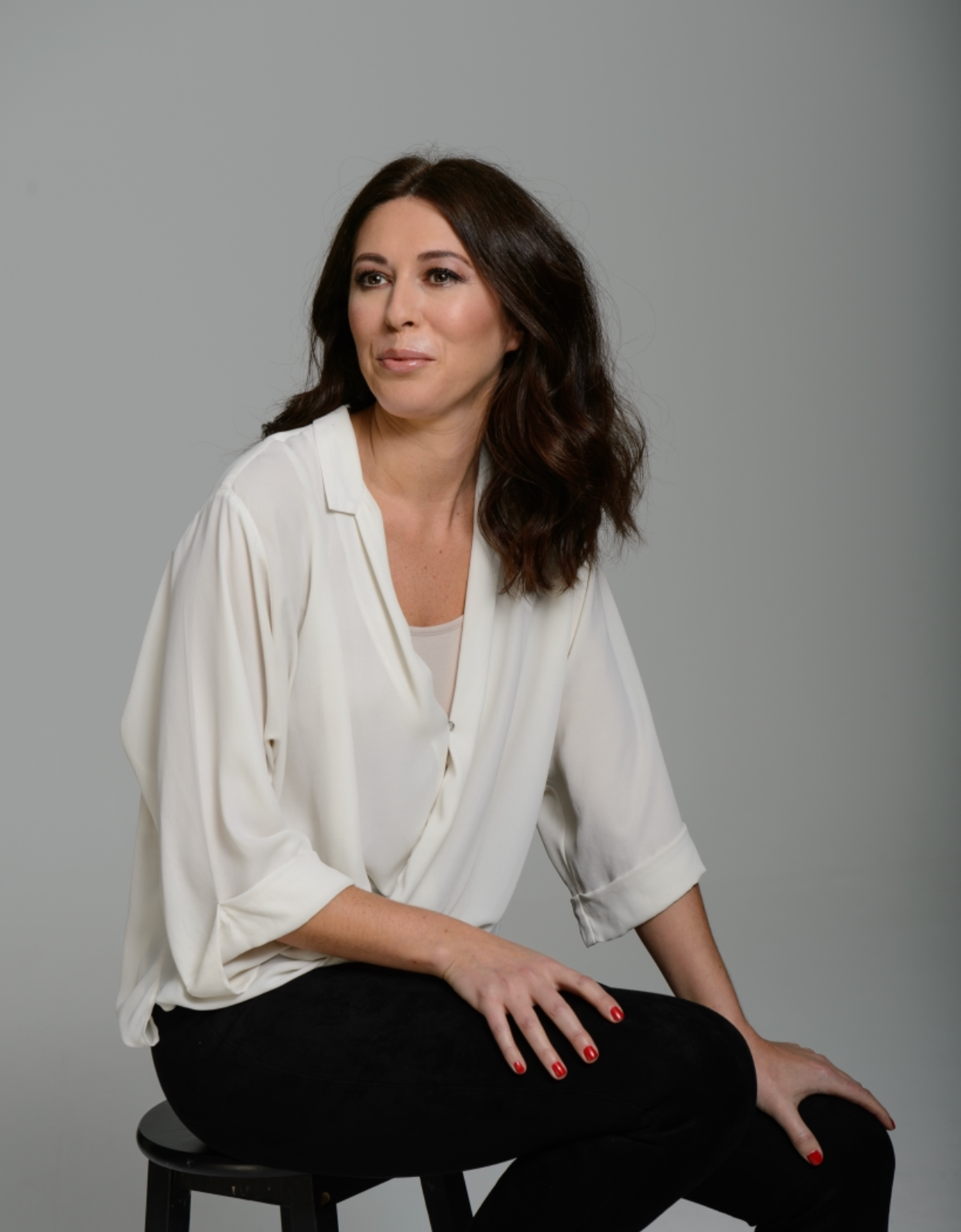 A woman with shoulder-length dark brown hair, wearing a white blouse and black pants, sitting on a black stool against a plain gray background.