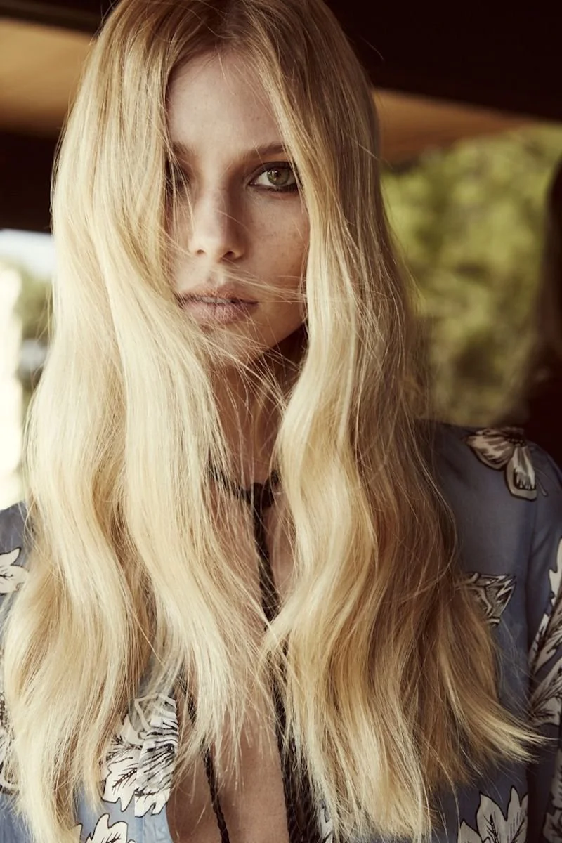 A young woman with long, wavy blonde hair and light skin looking directly at the camera, with a natural outdoor background.