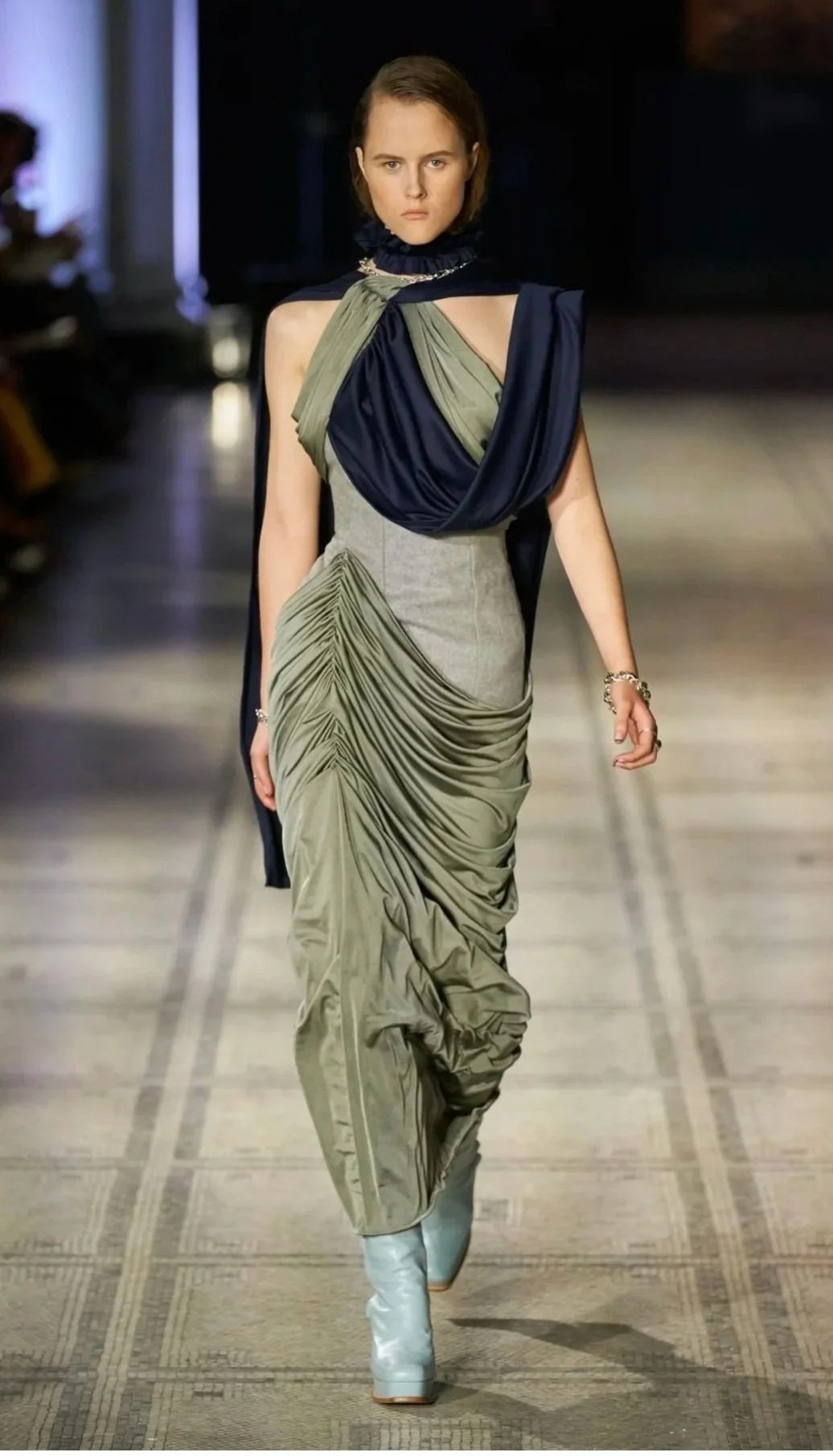Model walking on runway in a fashion show, wearing a designer dress with draped fabric in olive and navy colors, paired with white boots.
