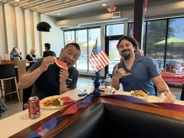 Two men sitting at a restaurant table with American flags centerpiece, enjoying a meal with watermelon and other food, in front of large windows showing a city view.