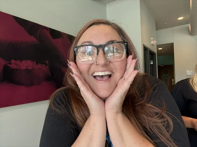 A woman with glasses smiling and holding her face with her hands in a cafe or restaurant.