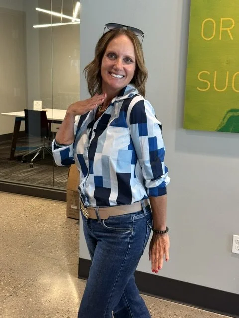 A woman with shoulder-length hair, wearing a blue, white, and navy patterned shirt, blue jeans, and glasses on her head, smiling and posing with one hand near her face in an indoor setting.