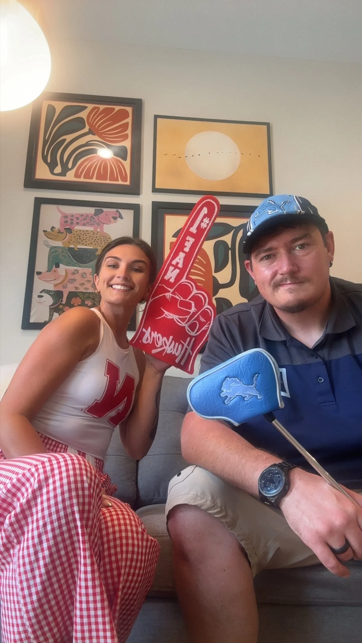 A woman and man sitting on a sofa in a room decorated with framed art, wearing casual clothes, holding foam fingers, and smiling for a selfie.