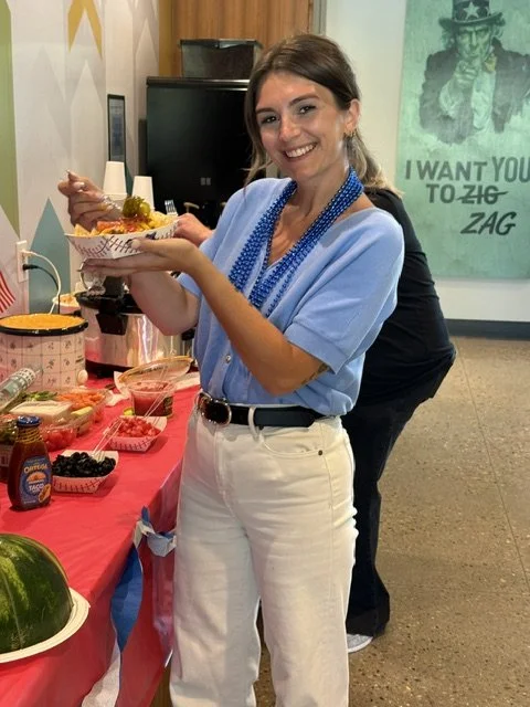 A woman standing at a table decorated for the 4th of July, smiling and holding a plate of food. The table has various snacks, a watermelon, and patriotic decorations.