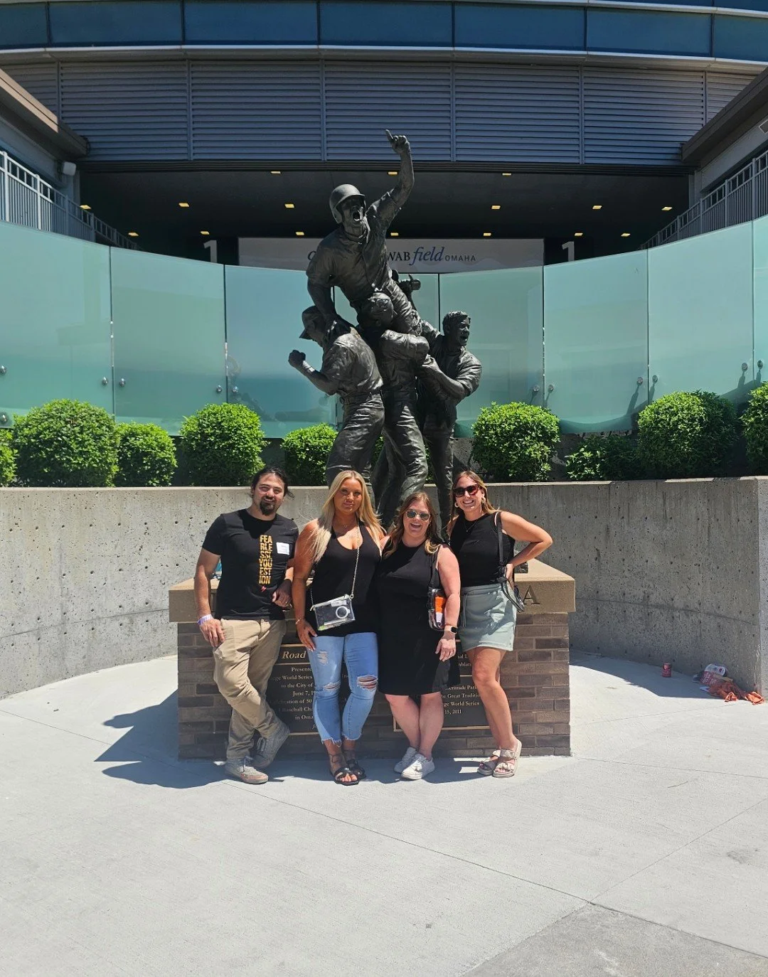 Four people posing in front of a monument with a large bronze statue depicting a military scene, with a modern building and greenery in the background.
