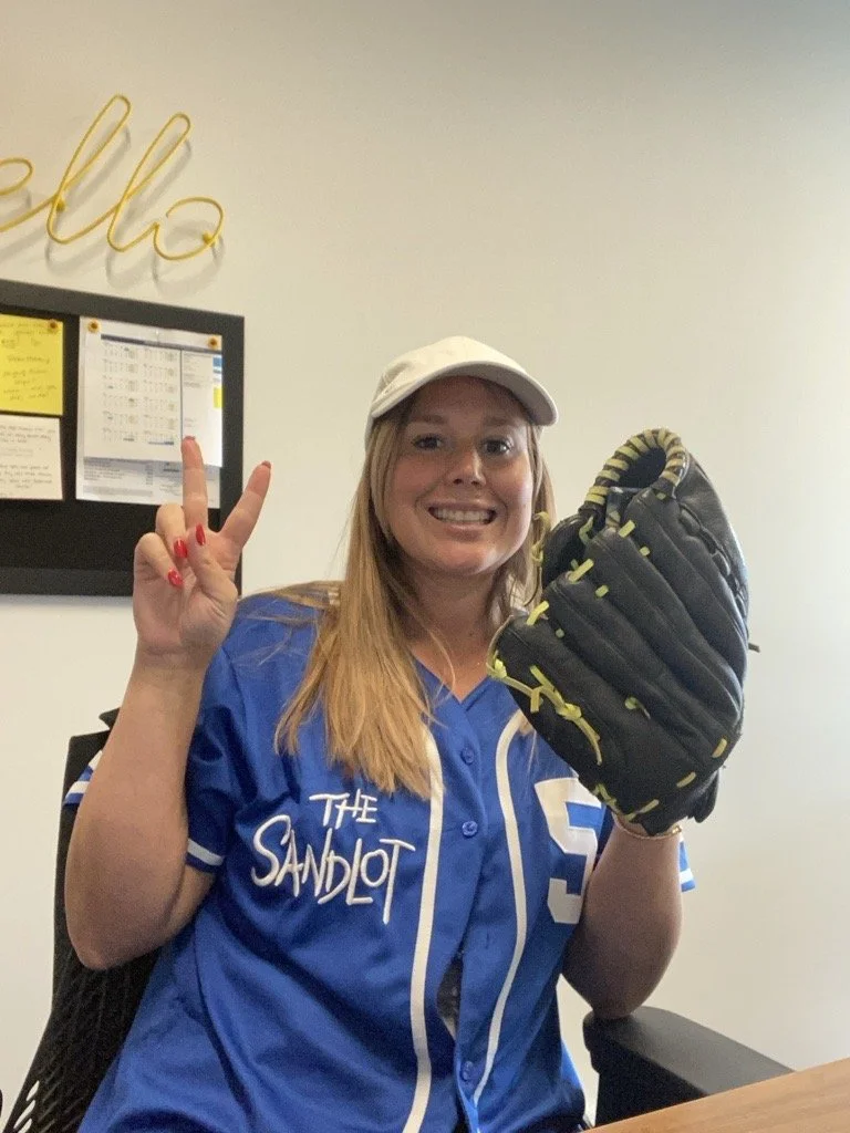 A woman wearing a blue sports jersey and a white cap holding a black and yellow baseball glove, smiling and making a peace sign indoors.
