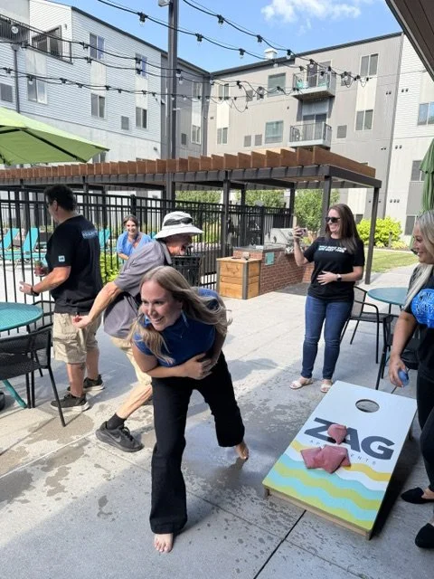 People playing beer pong outdoors on a sunny day, with some standing and others participating in the game, on a concrete patio with tables, chairs, and string lights overhead.