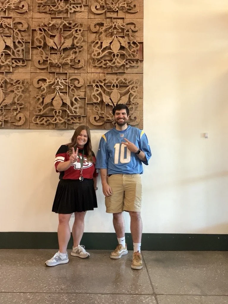 A young woman and a young man standing indoors against a decorative wooden wall. The woman is wearing a red and black sports jersey, a black skirt, and gray sneakers, and is making a peace sign with her fingers. The man is wearing a blue football jer