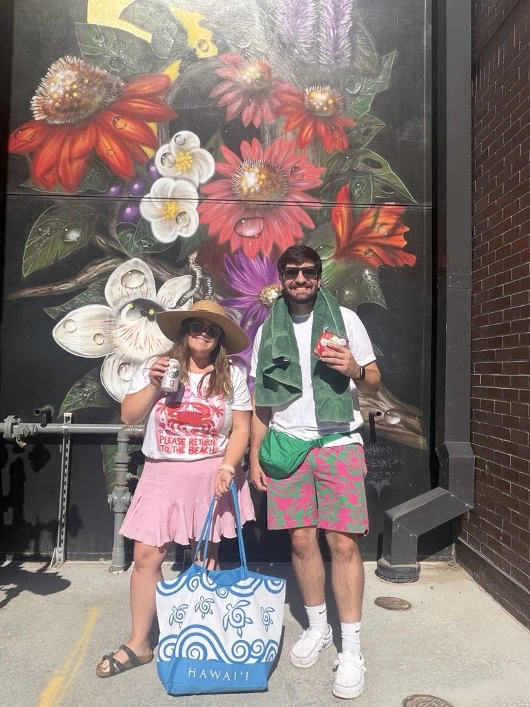 A man and a young girl standing in front of a colorful floral mural. The girl is holding a soda can and a tote bag labeled 'Hawaii,' wearing a pink skirt, white t-shirt, large sun hat, and sunglasses. The man is holding a drink, wearing sunglasses, a