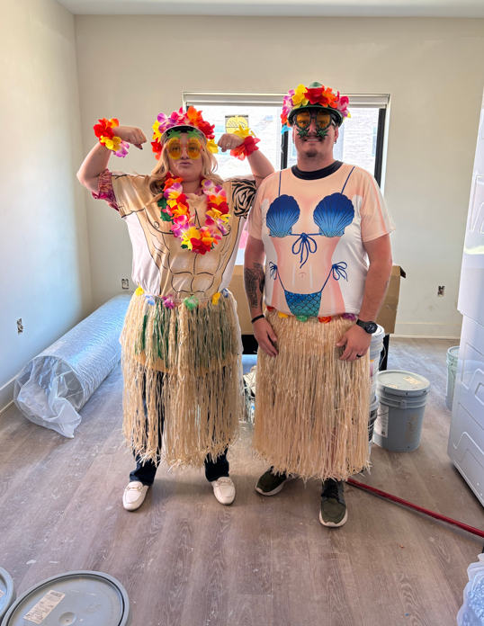 Two people dressed in hula costumes with grass skirts, floral leis, and flower crowns, standing in a room decorated for a tropical or Hawaiian-themed event.