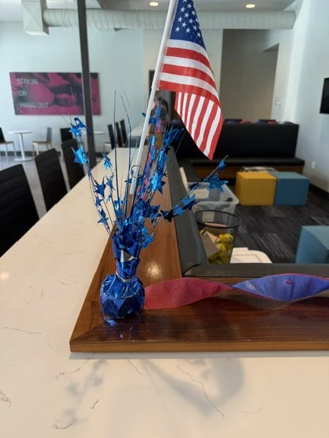 Small American flag on a table with blue star-shaped decorations in a blue vase, in a modern room with chairs and a TV in the background.