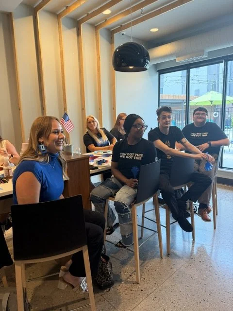 Group of young adults sitting at tables in a modern indoor space with large windows, some smiling and engaged, with a hanging black light fixture and American flags in the background.