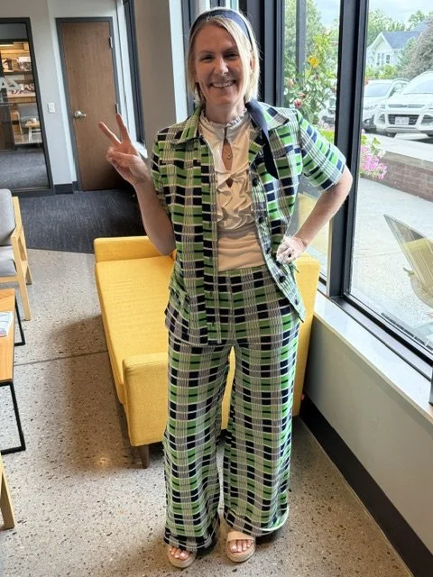 Woman indoors at a cafe or bookstore, wearing a green, black, and white plaid suit, making a peace sign with her right hand, smiling.