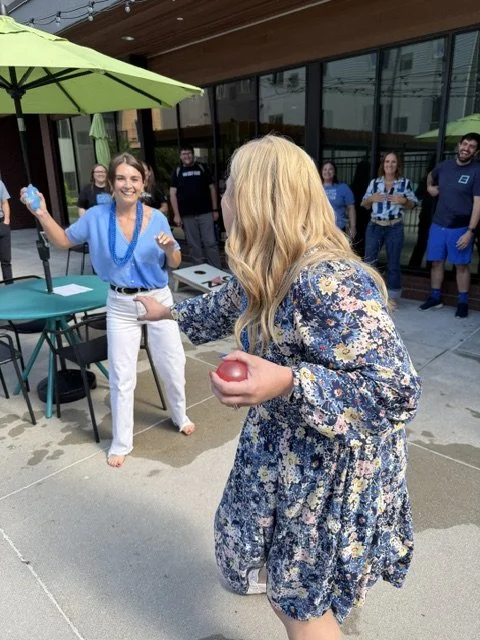 People playing a game with balls outside a building, with a woman in a floral dress holding a ball and smiling at another woman in a blue top and white pants.